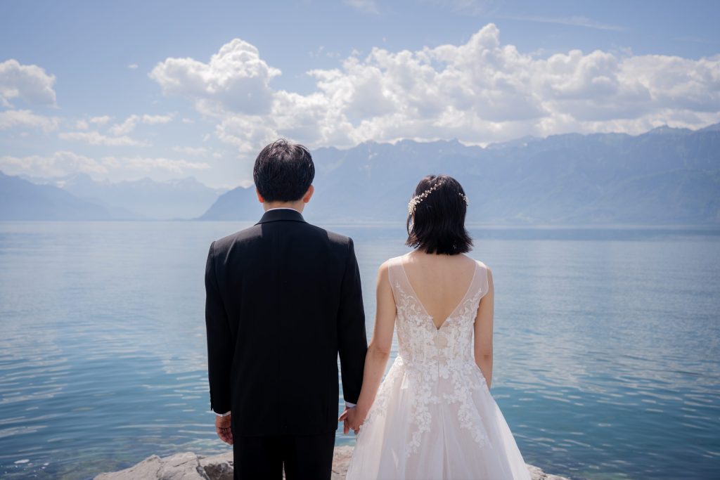 1 (87) Bride and groom seen from behind, hand in hand at the edge of Lake Geneva, mountains across the water