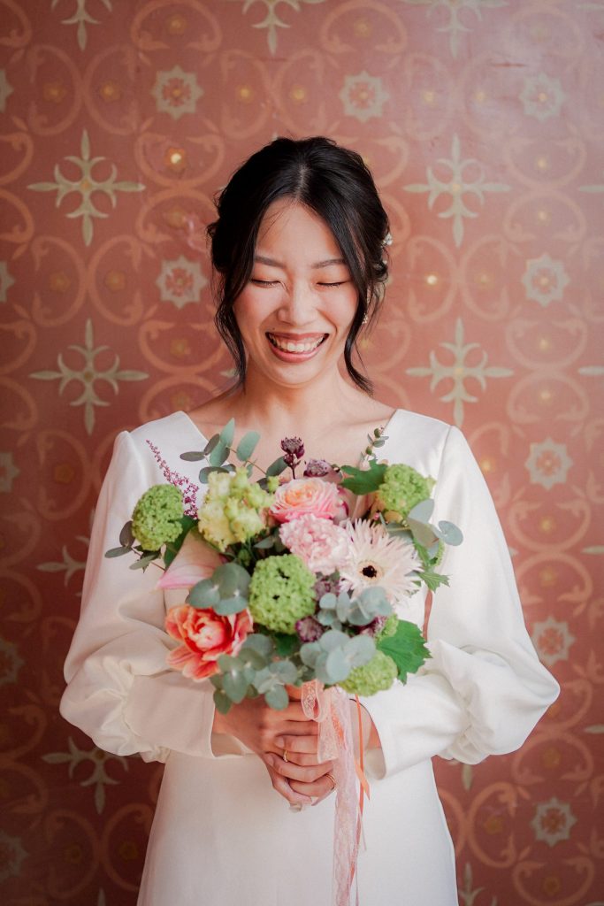 Vevey Wedding (61) Bride laughs with her eyes lowered, holding a bouquet of roses, hydrangea and eucalyptus, against a patterned terracotta wall