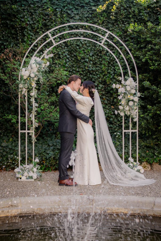 Vevey Wedding (96) Bride and groom embrace beneath a floral arch against a wall of ivy, her long veil trailing, fountain spray in the foreground