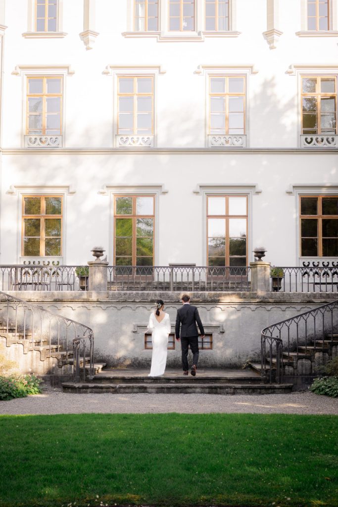 Vevey Wedding (99) Bride and groom walk up the stone steps towards a pale manor house, seen from the lawn below